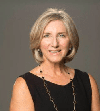 Anne Odette Kaye smiles at the camera, she is wearing a black dress, a long black and gold necklace, and pearl earrings.