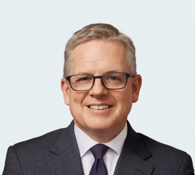 Neil McLaughlin smiles straight at the camera. He has short hair parted to one side, dark-rimmed glasses, and wears a dark grey suit with a dark tie.