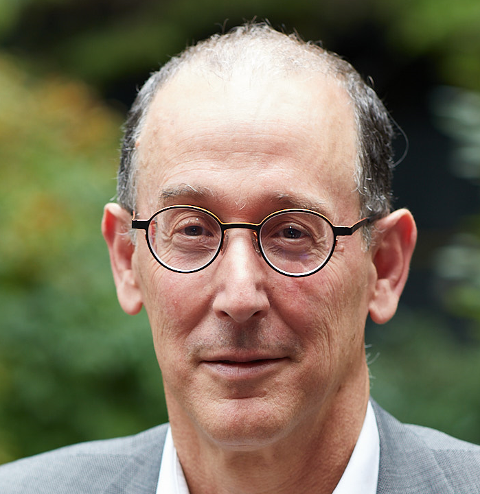Robert Beutel smiles at the camera in front of a background of greenery. He has short hair and wears round rimmed glasses with a light grey suit.