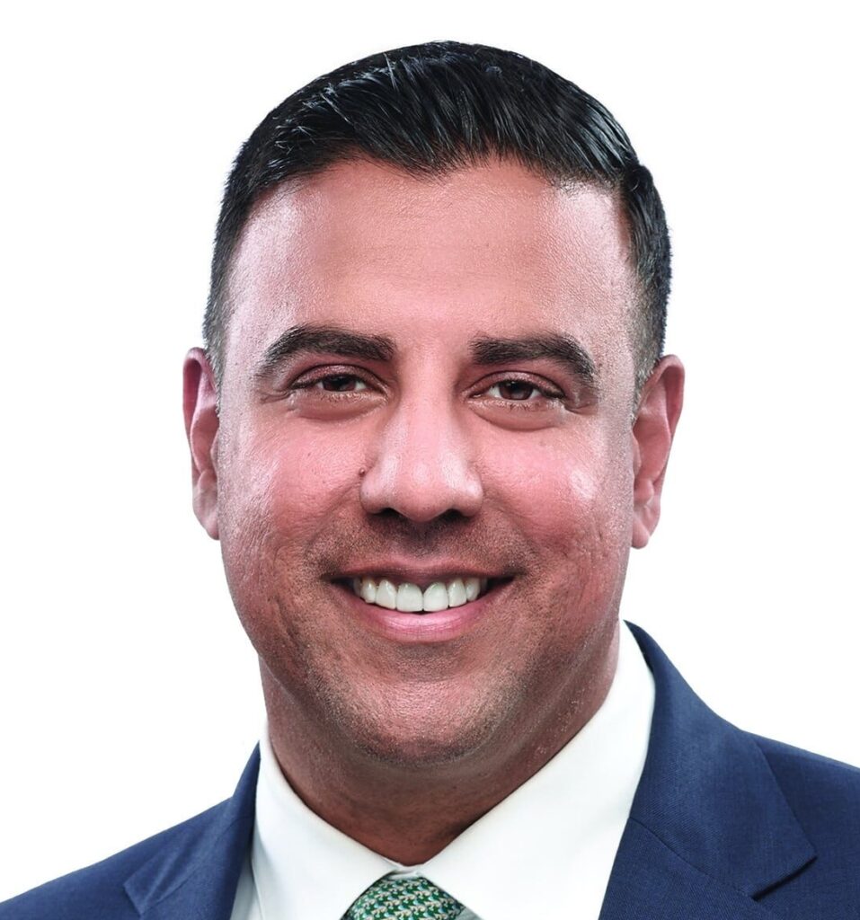 Ricco Bhasin smiles at the camera. He has short dark hair, brown eyes, and wears a dark blue suit with a green tie.