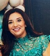 Umeeda Madhany smiles at the camera while leaning in for a photo. She has long dark hair and is wearing a teal dress in front of a bundle of balloons.