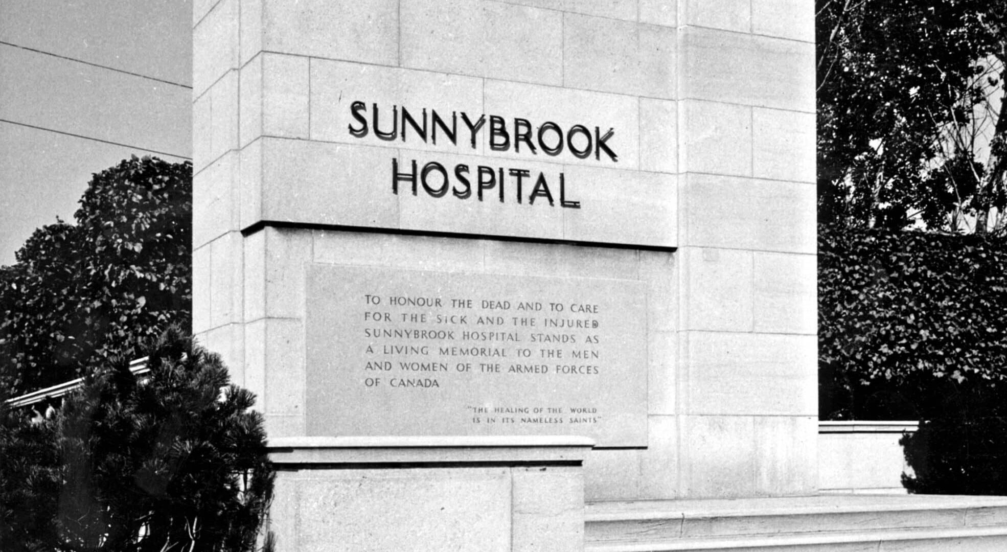 Black and white photo of the Sunnybrook Hospital memorial entrance sign, featuring the hospital's name engraved in stone beneath the Canadian coat of arms. Below the name is an inscription dedicating the hospital as a living memorial to the men and women of Canada’s armed forces. Trees and shrubs surround the stone monument.