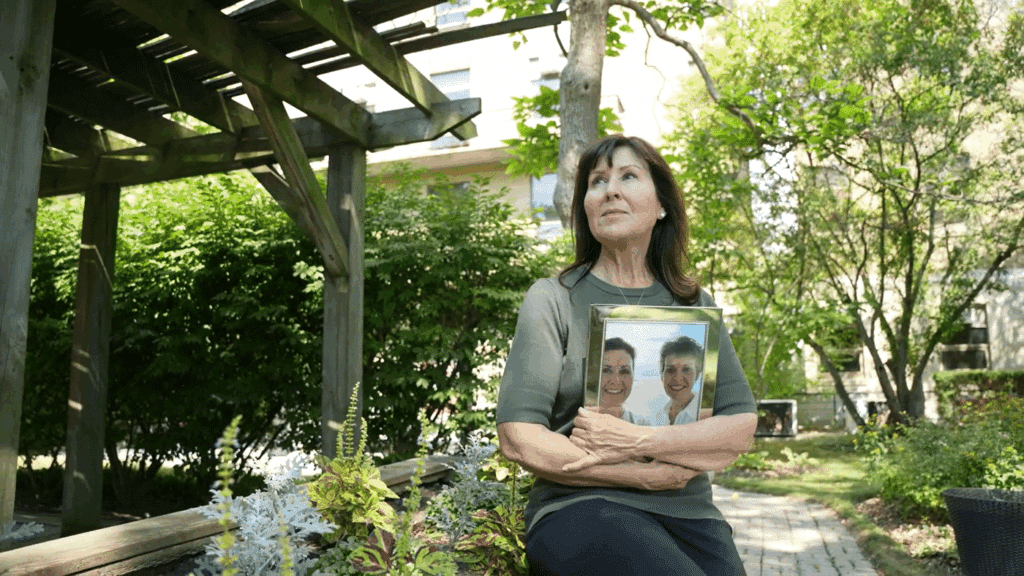 Woman sitting on a garden bench holding a framed photo of two people, with trees and a wooden structure in the background.